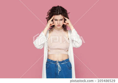 Portrait of confused puzzled woman with curly hair wearing casual style outfit thinking, keeps hands on temples, having serious expression. Indoor studio shot isolated on pink background. 105486529