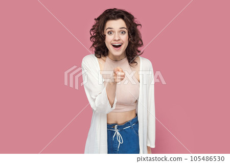 Portrait of amazed surprised woman with curly hairstyle wearing casual style outfit choosing you, looking at camera with big eyes and pleasant surprise. Indoor studio shot isolated on pink background. 105486530