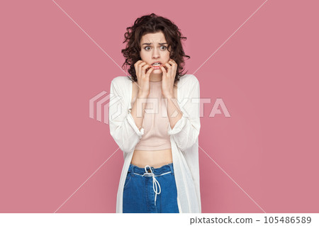 Portrait of nervous worried young adult woman with curly hair wearing casual style outfit, having troubles, biting her fingernails. Indoor studio shot isolated on pink background. Portrait of nervous worried young adult woman with curly hair wearing casual style outfit, having troubles, biting her fingernails. Indoor studio shot isolated on pink background. 105486589