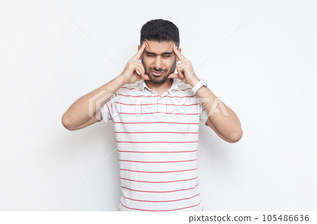 Portrait of sick unhealthy ill bearded man wearing striped t-shirt standing suffering headache, massaging his temples, frowning face. Indoor studio shot isolated on gray background. Portrait of sick unhealthy ill bearded man wearing striped t-shirt standing suffering headache, massaging his temples, frowning face. Indoor studio shot isolated on gray background. 105486636