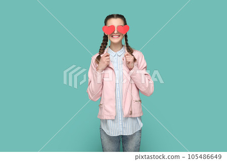 Portrait of romantic positive teenager girl with braids wearing pink jacket standing covering her eyes with two red hearts on stick. Indoor studio shot isolated on green background. 105486649