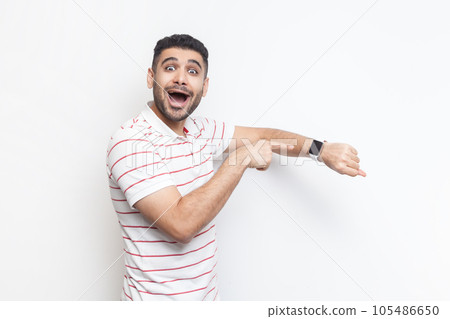 Portrait of amazed positive optimistic impressed bearded man wearing striped t-shirt standing looking at camera, pointing at smart watch. Indoor studio shot isolated on gray background. 105486650