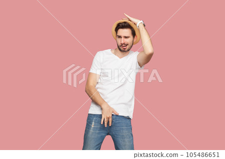 Portrait of excited playful handsome bearded man in white T-shirt and hat dancing, having funny pose, spending time on party. Indoor studio shot isolated on pink background. 105486651