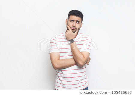 Portrait of pensive thoughtful attractive young adult bearded man wearing striped t-shirt standing holding chin, pondering, thinking. Indoor studio shot isolated on gray background. 105486653