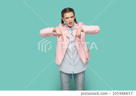 Portrait of strict bossy teenager girl with braids wearing pink jacket pointing both index fingers down, saying here and right now. Indoor studio shot isolated on green background. 105486657