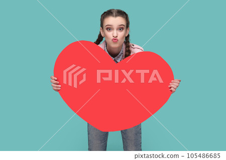 Portrait of optimistic romantic lovely teenager girl with braids wearing pink jacket holding big red heart, sending air kissing. Indoor studio shot isolated on green background. 105486685