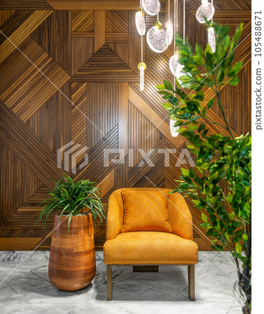 Modern orange armchair, tall rounded wooden planter with green bushes, tall glass chandelier, and wood cladding wall 105488671