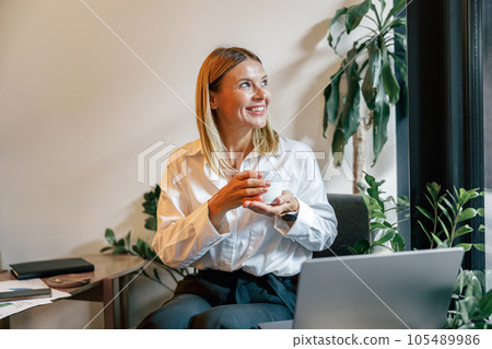 Smiling woman sales manager working on laptop and drinking coffee in coworking while looking at side 105489986
