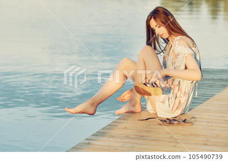 Young woman sits on edge of pier and takes off her sandals to soak her feet in water Young woman sits on edge of pier and takes off her sandals to soak her feet in water 105490739
