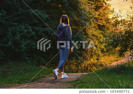 Back view of a woman goes along path from forest to clearing in rays of sunset 105490751