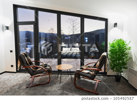 Light room with small coffee table and armchairs against large window with scenic view. Front view of living room with snowy mountain landscape through panoramic window at dusk. Concept of resting. 105492587