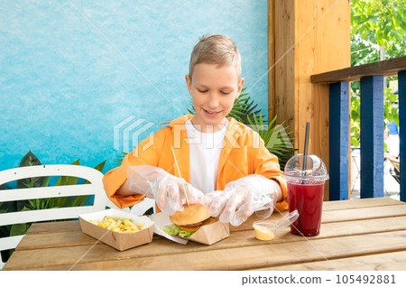 A cute boy in a street cafe eats fast food with lemonade in plastic disposable gloves 105492881