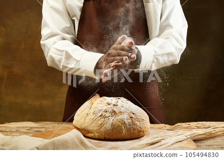Unrecognizable baker in bround apron pouring flour on round loaf of bread over brown background. Unrecognizable baker in bround apron pouring flour on round loaf of bread over brown background. 105494830