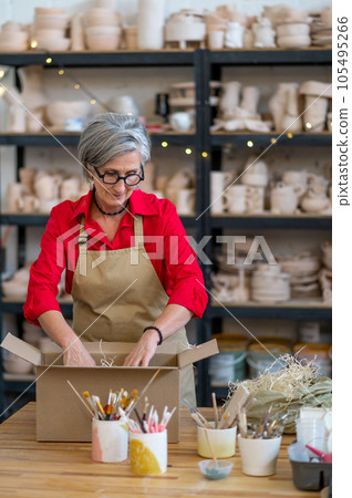 Female owner of small pottery shop standing by table and packing ordered item for clients. Female owner of small pottery shop standing by table and packing ordered item for clients. 105495266