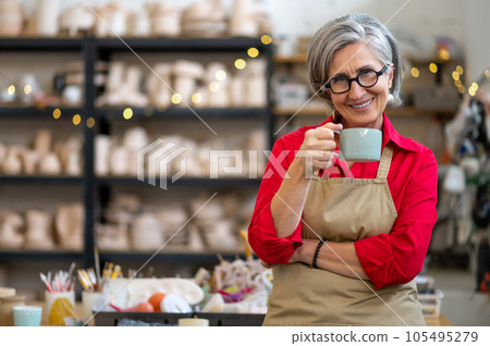 Happy mature female pottery master taking coffee break in ceramics studio. Happy mature female pottery master taking coffee break in ceramics studio. 105495279