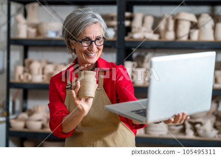Woman pottery artist using laptop in art studio showing her earthenware products, selling online. 105495311