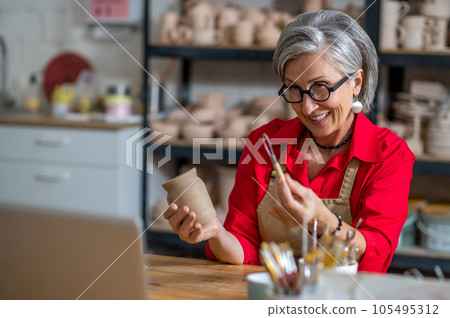 Senior woman potter broadcasting live lesson masterclass painting clay products, teaching students online. Senior woman potter broadcasting live lesson masterclass painting clay products, teaching students online. 105495312