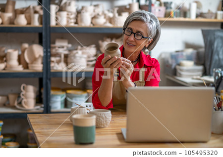 Mature woman painting pottery earthenware product in front of laptop while live streaming. Mature woman painting pottery earthenware product in front of laptop while live streaming. 105495320