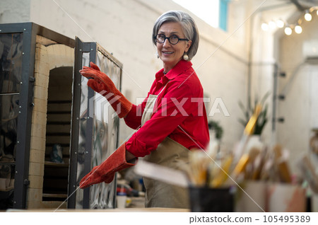 Craftswoman placing clay mugs into kiln for firing pottery while working in workshop. Craftswoman placing clay mugs into kiln for firing pottery while working in workshop. 105495389