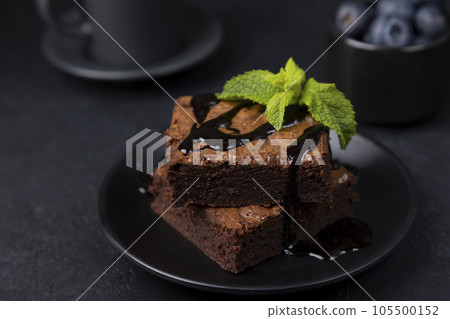 Chocolate brownie cake with mint, blueberries, chocolate topping and espresso. Traditional American dessert. Popular cake with dark chocolate on a black background. Close-up, selective focus. 105500152