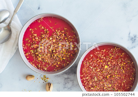 Malabi traditional Arabic dessert. Milk pudding with pink syrup and pistachio in gray bowl, white marble background. Malabi traditional Arabic dessert. Milk pudding with pink syrup and pistachio in gray bowl, white marble background. 105502744