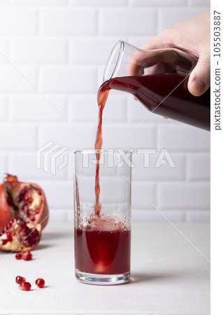 A man pours pomegranate juice from a bottle into a tall glass with ice. The concept of freshness and usefulness. In the background, the fruit and seeds of a pomegranate. Selective focus, close-up. A man pours pomegranate juice from a bottle into a tall glass with ice. The concept of freshness and usefulness. In the background, the fruit and seeds of a pomegranate. Selective focus, close-up. 105503887
