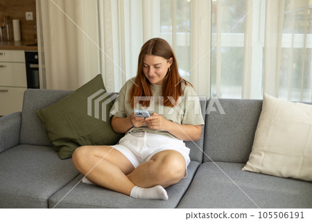 Casual beautiful woman is using phone sitting on a sofa. Phone Communication. 30s gorl laughing holding smartphone, enjoying doing online ecommerce shopping in mobile apps or watching video Casual beautiful woman is using phone sitting on a sofa. Phone Communication. 30s gorl laughing holding smartphone, enjoying doing online ecommerce shopping in mobile apps or watching video 105506191
