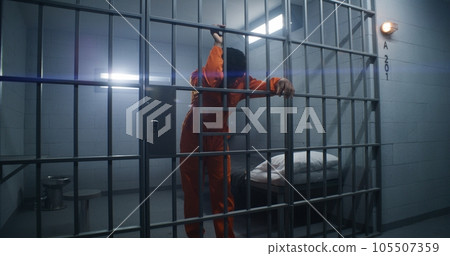 Prisoner in orange uniform leans on jail cell bars 105507359