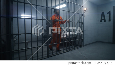 Prisoner in orange uniform leans on jail cell bars 105507382