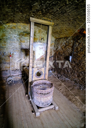 There is little light in the dark basement room. The guillotine, a medieval tool of punishment, stands with a basket made of wicker There is little light in the dark basement room. The guillotine, a medieval tool of punishment, stands with a basket made of wicker 105508035
