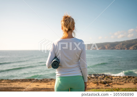 Senior woman holding yoga mat on ocean shore back view 105508890