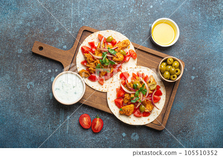 Cooking Traditional Greek Dish Gyros: Pita bread with vegetables, meat, herbs, olives on rustic wooden cutting board with Tzatziki sauce, olive oil from above on dark blue stone background. Cooking Traditional Greek Dish Gyros: Pita bread with vegetables, meat, herbs, olives on rustic wooden cutting board with Tzatziki sauce, olive oil from above on dark blue stone background. 105510352