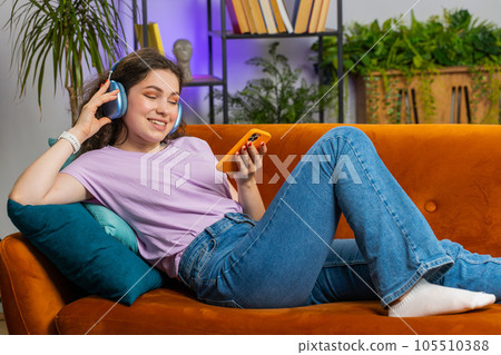 Happy young woman in wireless headphones relaxing lying on sofa at home listening favorite music 105510388