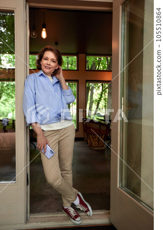 Mature woman in casual clothes on the porch of country cottage. Attractive middle-aged brunette with cell phone in her hand. Senior caucasian lady uses smartphone. Active lifestyle for the elderly. 105510864