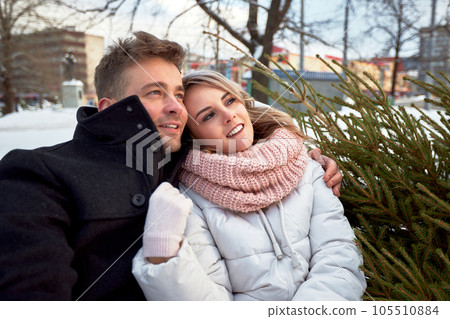Happy couple in love in Christmas market. Young beautiful man and woman in winterwear embracing and looking dreamily into the distance. Romantic outside dating in winter snowy city. Happy couple in love in Christmas market. Young beautiful man and woman in winterwear embracing and looking dreamily into the distance. Romantic outside dating in winter snowy city. 105510884