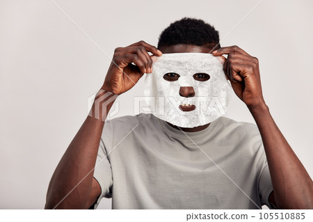 Studio portrait of young black man applying moisturizing paper mask on his face. Happy African American millennial man uses beauty products in his skincare routine to maintain healthy look. Studio portrait of young black man applying moisturizing paper mask on his face. Happy African American millennial man uses beauty products in his skincare routine to maintain healthy look. 105510885