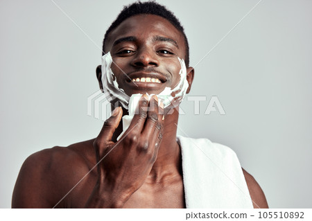 Studio portrait of handsome black guy with naked torso applying cream on his face. Afro American man with towel on his shoulder uses beauty products in his skincare routine to keep his healthy look. 105510892