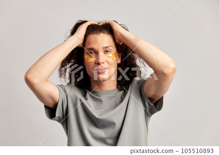 Young handsome long-haired guy applying under eye hydrogel patches on his face. Caucasian brown-haired millennial man practices skin care routine to keep healthy and youthful looking. Studio portrait. Young handsome long-haired guy applying under eye hydrogel patches on his face. Caucasian brown-haired millennial man practices skin care routine to keep healthy and youthful looking. Studio portrait. 105510893