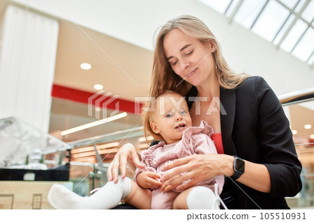 Young pretty mother with a small baby sitting on a bench in a shopping center. Mom and daughter in pink clothes hug, play, relax and have fun while shopping. Family weekend in shopping mall. 105510931