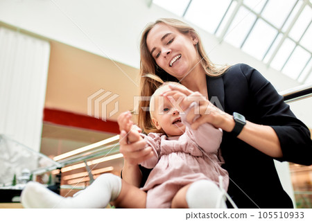 Young pretty mother with a small baby sitting on a bench in a shopping center. Mom and daughter in pink clothes hug, play, relax and have fun while shopping. Family weekend in shopping mall. 105510933