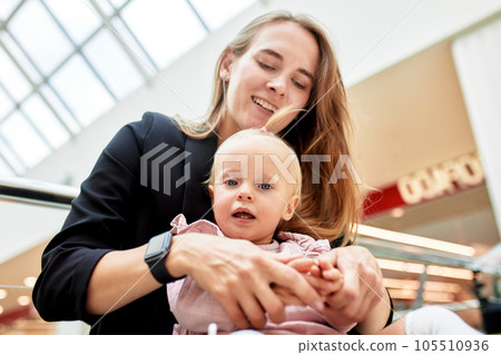 Young pretty mother with a small baby sitting on a bench in a shopping center. Mom and daughter in pink clothes hug, play, relax and have fun while shopping. Family weekend in shopping mall. 105510936