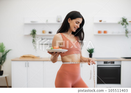 Happy woman measuring waist with tape after workout at home Happy woman measuring waist with tape after workout at home 105511403