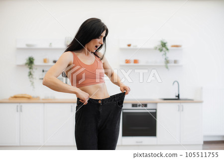 Woman looking at oversized jeans on thin waist at home Woman looking at oversized jeans on thin waist at home 105511755