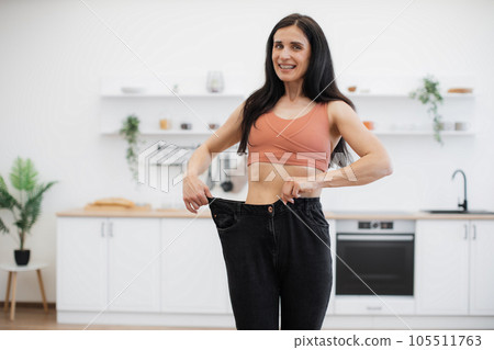Woman looking at oversized jeans on thin waist at home 105511763