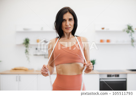 Woman in gym clothes posing with measuring tape in kitchen 105511793