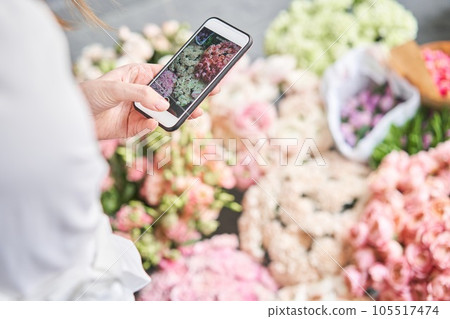 A woman takes a photo on her phone of flowers. Floral shop concept . Florist woman creates flower arrangement in a wicker basket. . Flowers delivery. A woman writes a video blog or online course A woman takes a photo on her phone of flowers. Floral shop concept . Florist woman creates flower arrangement in a wicker basket. . Flowers delivery. A woman writes a video blog or online course 105517474