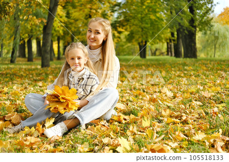 The family has fun in the park. Girl and mother sit on the ground among yellow leaves, smile and look at the camera 105518133