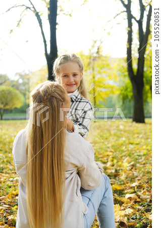 Mother and daughter spend time together in the autumn park. Daughter holding mother's hands, smiling and looking at camera Mother and daughter spend time together in the autumn park. Daughter holding mother's hands, smiling and looking at camera 105520851