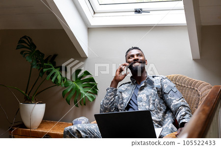 African american male soldier wearing military uniform with laptop and talking on smartphone at home 105522433
