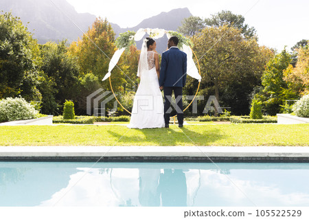 Rear view of african american bride and groom under wedding arch by pool in sunny garden 105522529